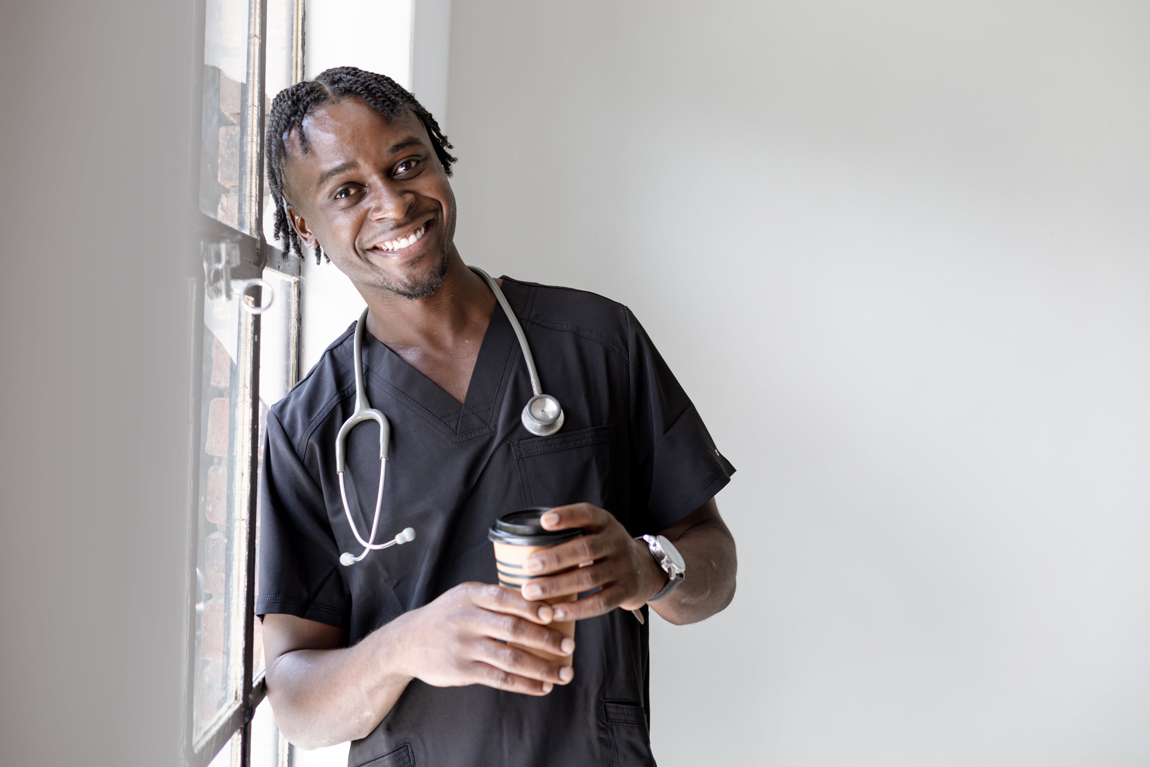 Doctor in black scrubs holding a coffee cup, standing by a window.